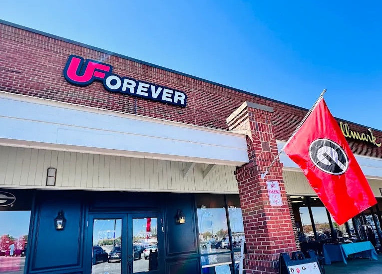 UF Forever restaurant storefront with brick facade, black double doors, a red flag, and a sidewalk sign on a clear blue sky day.